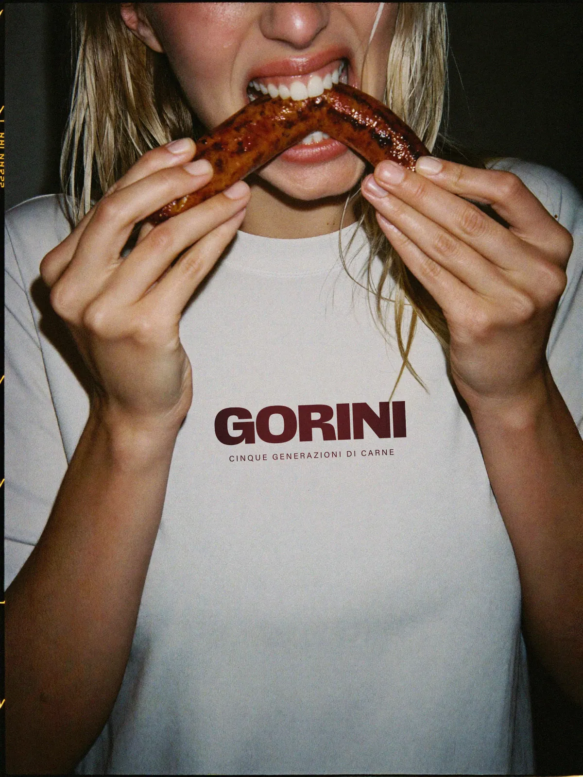 Ragazza in t-shirt Gorini Cinque generazioni di carne che morde una salsiccia alla griglia, foto con flash diretto per il rebranding Gorini Carne