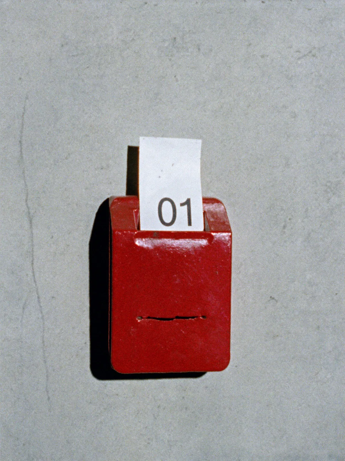 Distributore rosso di biglietti eliminacode su un muro di cemento con il biglietto numero 01: HABA Studio lavora a numero chiuso