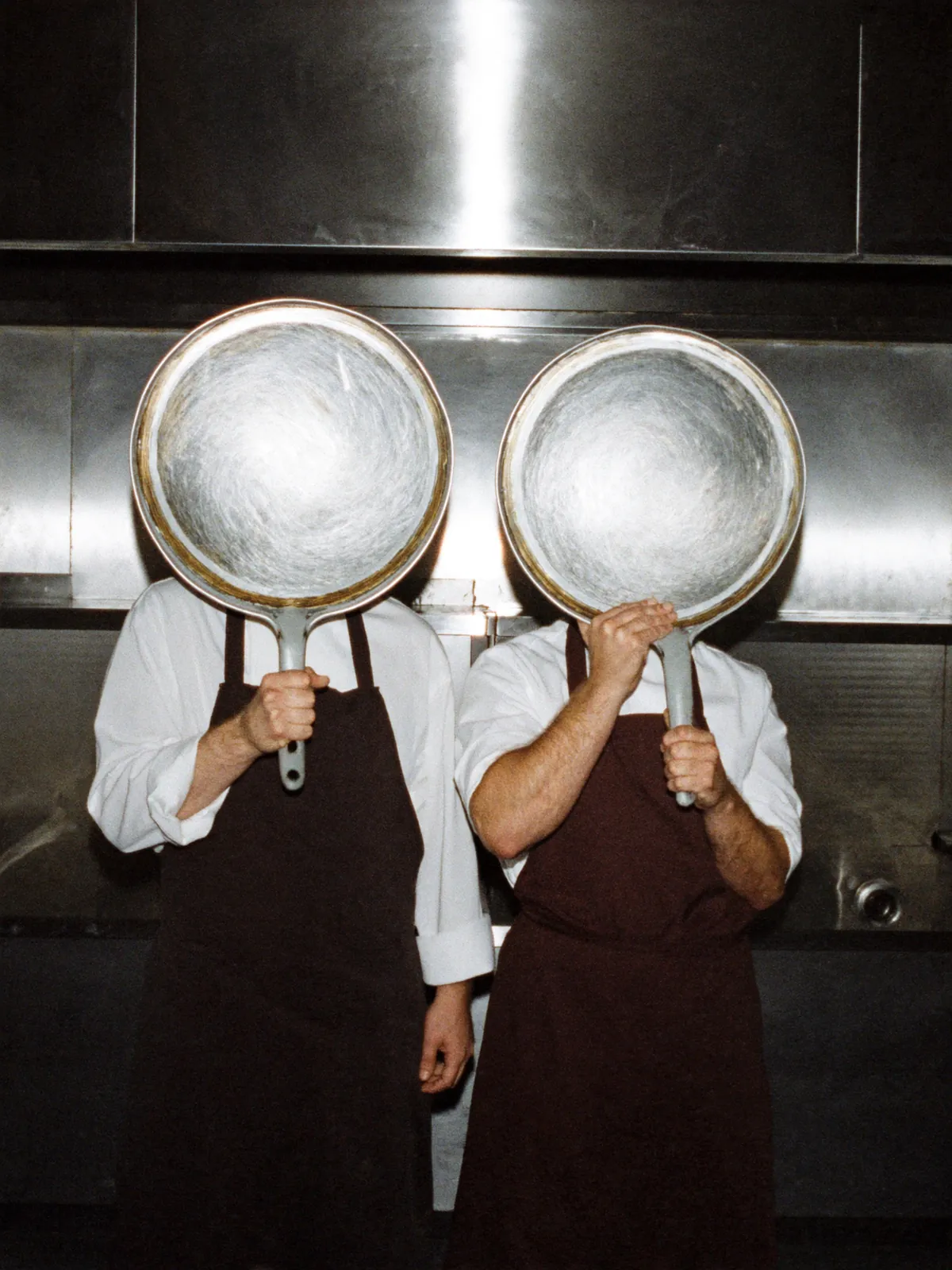 Due cuochi in grembiule che si coprono il volto con due grandi padelle in una cucina inox, foto con flash per OSTIA Cucina